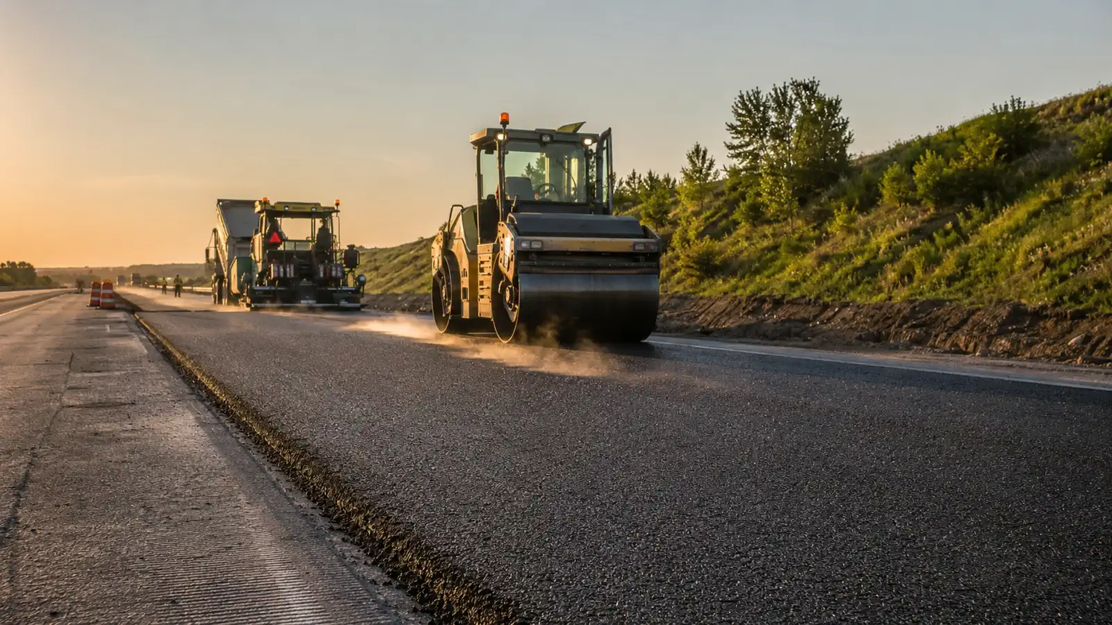 Asphalt paver laying a fresh mat of hot mix on a highway at golden hour with a steel drum roller compacting behind it